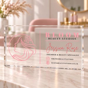May include: A rectangular clear acrylic sign featuring a pink logo of a woman's profile with a flower, the text BLOOM BEAUTY STUDIOS, the name Jessica Rose, the title FOUNDER & BEAUTY SPECIALIST, and social media handles @BLOOMBEAUTYSTUDIO.
