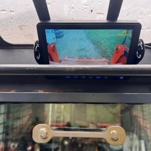 May include: A black monitor displaying a view from a vehicle's rear. The screen shows a dirt path and orange machinery. The monitor is mounted on a dark surface, with a metal bar and a yellow vehicle visible below.