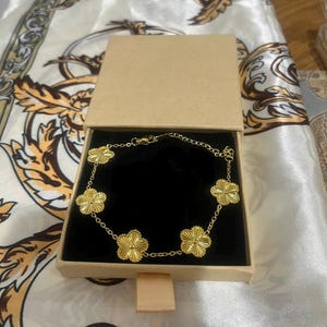 May include: A gold-colored bracelet with flower-shaped charms, displayed in an open, tan jewelry box with a black interior. The bracelet is on a white and gold patterned fabric.
