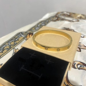 May include: A gold-colored bangle bracelet with decorative circular accents is displayed on a beige box with a black interior. The bracelet is circular and has a clasp. The background is a patterned fabric.