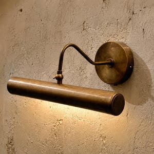 May include: A wall-mounted picture light with an antique brass finish. The fixture features a circular wall plate, a curved arm, and a horizontal cylindrical lamp head that emits a warm downward glow from its underside, illuminating the wall.