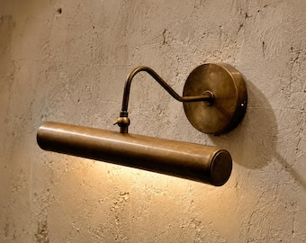 May include: A wall-mounted picture light fixture with an antique brass finish. The lamp features a cylindrical horizontal shade attached to a circular wall plate by a curved, adjustable arm, emitting a warm downward glow.