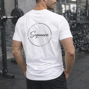 Puede incluir: Camiseta blanca de cuello redondo con la palabra "Sequence" en un diseño circular en la espalda. El texto "SYSTEM OF ATHLETIC" y "BUILT BY SCIENCE" también están en el diseño. La persona lleva pantalones cortos negros.