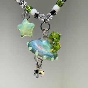 May include: A beaded necklace featuring a clear star charm, a glass UFO with a green cat figure, and a small black and white cow charm. The necklace is strung with clear, white, black, and green beads on a silver-toned chain.