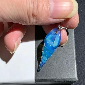 Peut inclure: Un pendentif en verre bleu et turquoise en forme de coquillage en spirale, suspendu à une chaîne argentée. Le pendentif est translucide, avec des couleurs tourbillonnantes et une finition brillante. Le bijou est présenté sur un fond sombre.