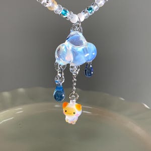 May include: A necklace featuring a light blue glass cloud pendant with dangling dark blue glass raindrops and a small orange and white glass creature charm. The chain is composed of clear, white, and blue beads with silver-toned metal accents.