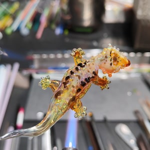 Puede incluir: Una figura de gecko de vidrio hecha a mano con un cuerpo translúcido de color ámbar y amarillo, manchas de color marrón oscuro y una superficie texturizada y rugosa. La escultura con forma de lagarto se sostiene sobre una llama azul.