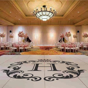 May include: A white dance floor with a black monogram design featuring a large letter "H" in a decorative frame. The dance floor is in a large ballroom with white drapes and floral centerpieces on the tables.