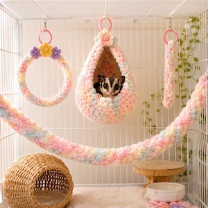 May include: A sugar glider sits inside a pastel-coloured, crocheted teardrop-shaped hanging pouch. The set includes a matching crocheted ring, a long hanging rope, and a woven wicker dome-shaped hideout, all arranged within a wire cage enclosure.