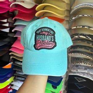 Peut inclure: Une casquette de baseball bleu clair avec un écusson brodé noir sur le devant portant le texte "THAT SOUNDS LIKE my HUSBAND'S Problem" en lettres blanches et roses, entourée d'une pile de diverses casquettes de baseball colorées en arrière-plan.