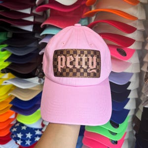 May include: A light pink baseball cap featuring a rectangular patch on the front. The patch has a brown and black checkered pattern with the word petty written in a stylized, light pink gothic-inspired font across the center of the design.