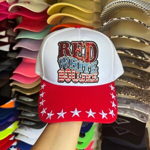 May include: A white baseball cap with a red brim featuring white embroidered stars along the edge. The front panel displays the text RED WHITE BOUJEE in red and blue glittery lettering with a patriotic striped pattern inside the letters.