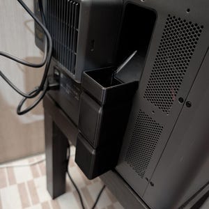 May include: A black 3D printed waste chute attachment mounted to the side of a dark grey 3D printer. The rectangular plastic accessory features a vertical opening and is positioned directly below the printer's rear exhaust vent and cooling fan.