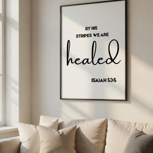 May include: A framed print with the words "BY HIS STRIPES WE ARE healed" in black script. The text is set against a white background, with the Bible verse Isaiah 53:5 below. The print hangs above a beige sofa with several cushions.