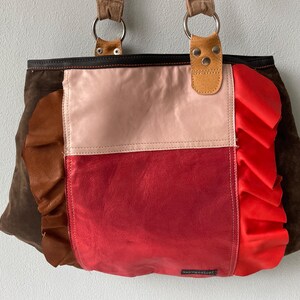 Op de afbeelding: Een patchwork leren tote bag met een mix van kleuren. De tas heeft een rood, beige en bruin kleurenschema met een gerimpelde rand aan de rechterkant. De tas heeft twee bruine handvatten die met zilveren ringen zijn bevestigd.
