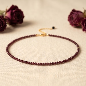 Op de afbeelding: Een delicate ketting met kleine, ronde, dieprode kralen. De ketting heeft een goudkleurige ketting en sluiting. Gedroogde bordeauxrode rozen op de achtergrond voegen een vleugje elegantie toe aan de afbeelding.
