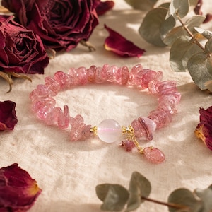 Pode incluir: Uma pulseira composta por contas irregulares e translúcidas de quartzo morango, com uma única conta lisa, redonda e rosa pálido no centro. O design inclui espaçadores de metal dourado e um pequeno pingente de pedra rosa pendurado.