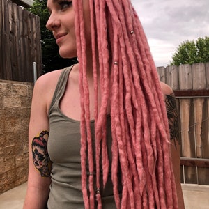 May include: A person with long, pink dreadlocks. The dreadlocks are tied with silver rings. The person is wearing a green tank top and black jeans. They have a tattoo on their arm of a cartoon face with red eyes.