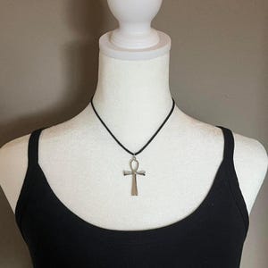 May include: A silver-colored ankh pendant necklace on a black cord. The ankh is a symbol of life and is suspended from a black cord. The necklace is displayed on a white mannequin bust, with a black tank top.