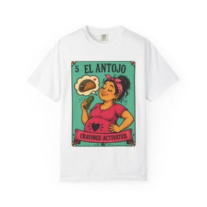 May include: White t-shirt featuring a colorful illustration. The graphic shows a woman holding a pickle, with a thought bubble of a taco. Text includes "5 EL ANTOJO" and "Cravings Activated."