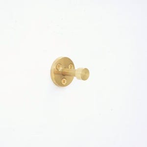 May include: A circular brass wall hook with a cylindrical peg and a flat, rounded end. The base plate features three visible screw holes with matching brass screws, providing a secure mounting point for the hardware against a flat surface.