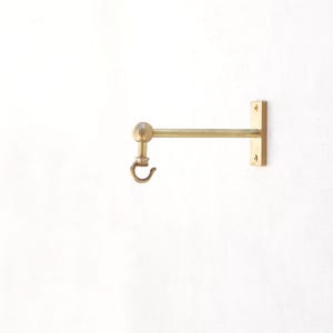 May include: A brass-toned wall hook with a rounded ball detail and a curved hook. The hook is mounted on a rectangular plate with two screw holes. The design is simple and functional, suitable for hanging items.