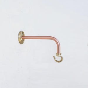 May include: A wall-mounted hook featuring a horizontal copper-toned arm extending from a circular brass mounting plate with three screw holes. The arm curves downward at the end, terminating in a small, polished brass hook for hanging items.
