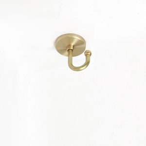 May include: A wall-mounted hook with a circular base plate and a curved arm ending in a small spherical knob. The hardware features a brushed gold-tone metallic finish with a smooth, uniform texture throughout the entire structure of the hook.