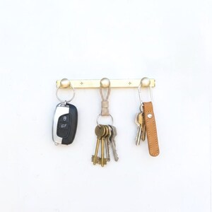 May include: A brass key holder with three key rings, each holding different keys. The key holder is mounted on a white wall. One key ring holds a car key, another holds a bundle of keys, and the third holds keys with a tan leather fob.