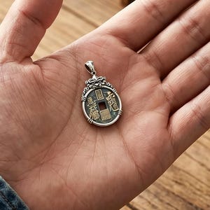 Puede incluir: Un colgante de plata con un diseño de moneda antigua, sostenido en una mano. El colgante presenta un centro cuadrado con caracteres chinos y detalles ornamentados en los bordes. El fondo incluye una superficie de madera y una bolsa de cuero.