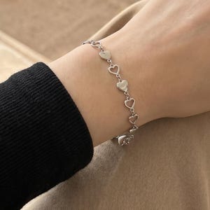May include: A silver-toned chain bracelet featuring a series of small, hollow heart-shaped links connected in a continuous row. The bracelet is crafted from polished metal and rests against the wrist, showcasing a delicate and uniform design.