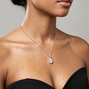 Peut inclure: Un collier en argent avec un pendentif en forme de poire. Le pendentif est une pierre précieuse transparente sertie dans un cadre argenté. Le collier est porté sur un haut noir sans bretelles.