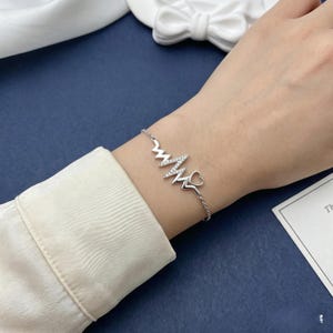 Op de afbeelding: Een zilveren armband met een hartslagontwerp en een klein hartje aan het einde. Het hartslaggedeelte is versierd met kleine, sprankelende steentjes. De armband wordt om een pols gedragen, wat het delicate en elegante ontwerp benadrukt.