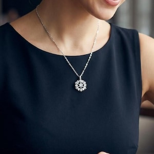 Könnte beinhalten: Eine silberne Halskette mit einem Schneeflocken-Anhänger. Der Anhänger hat ein detailliertes Schneeflocken-Design mit kleinen, funkelnden Akzenten. Die Halskette wird auf einem schwarzen Oberteil getragen, was einen Kontrast erzeugt, der den Schmuck hervorhebt.