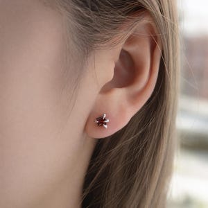 May include: A small stud earring featuring a deep red, star-shaped gemstone held in place by four silver-toned prongs. The earring is positioned on the earlobe, showcasing a metallic setting that secures the dark red stone in a star-like configuration.