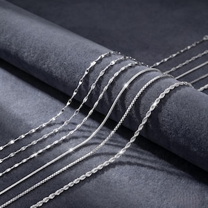 Peut inclure: Plusieurs colliers en argent présentés sur un fond de velours gris foncé. L'image présente différents styles de chaînes, notamment des modèles à câble, à maillons carrés et torsadés. Les colliers sont brillants et réfléchissants.