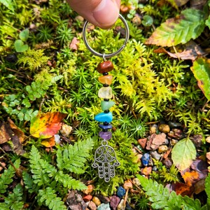 May include: A keychain featuring a series of colorful gemstones in a chakra arrangement, topped with a silver ring and a Hamsa hand charm. The gemstones include brown, orange, yellow, green, blue, indigo, and purple. The background is a bed of green moss and foliage.