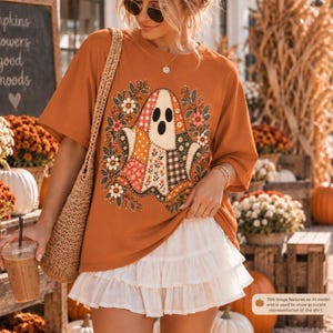 May include: An orange short-sleeve t-shirt featuring a central graphic of a ghost made of various patchwork patterns, surrounded by a floral wreath. The shirt is paired with a white tiered mini skirt and a woven shoulder bag.