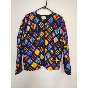 May include: A long-sleeved jacket featuring a black grid pattern filled with multicolored woven ribbon squares in shades of yellow, blue, purple, and red. The garment has a round neckline, a front opening, and a white label at the inner collar.