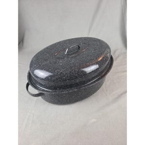 May include: An oval-shaped roasting pan with a matching lid, featuring a dark gray enamel finish with white speckles. The pan includes two side handles and a small handle on the lid, providing a functional design for cooking and food preparation.