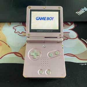 Op de afbeelding: Een roze Game Boy Advance SP handheld gameconsole, opengeklapt om het "GAME BOY" logo op het scherm te tonen. De console heeft een kruisje, A- en B-knoppen en start/select knoppen. Het scherm wordt omlijst door een zwarte rand.