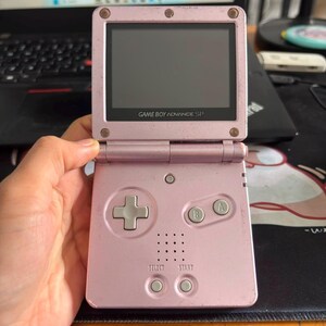 Puede incluir: Una consola de videojuegos portátil Game Boy Advance SP de color rosa claro con pantalla abatible, cruceta direccional, botones A y B, y botones Select y Start. El dispositivo tiene un acabado metálico con signos visibles de desgaste.