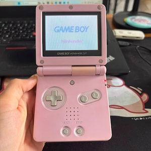 Puede incluir: Una consola de juegos portátil Game Boy Advance SP de color rosa claro con diseño plegable. La pantalla muestra el texto GAME BOY y Nintendo. El dispositivo cuenta con una cruceta, botones A y B, y botones Select y Start plateados.