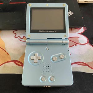 May include: A light blue Game Boy Advance SP handheld gaming console with a flip-top design. The device features a square screen, a directional pad, A and B buttons, Select and Start buttons, and a speaker grille. The text Game Boy Advance SP is printed below the screen.
