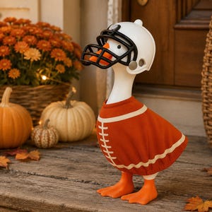 Könnte beinhalten: Eine weiße Gänsefigur, die einen weißen Football-Helm mit schwarzem Gesichtsschutz und ein orangefarbenes Trikot mit weißen Streifen und Schnürsenkeln trägt. Die Gans hat leuchtend orangefarbene Schwimmfüße und einen passenden Schnabel.