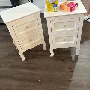 May include: Two white bedside tables, each featuring two drawers with clear, round knobs. The tables have a rectangular top surface and curved legs. The design includes recessed drawer panels, providing a clean, uniform appearance for these storage units.
