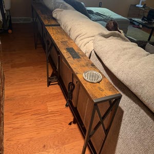 May include: A long, narrow sofa table with a rustic brown wood-grain finish and a black metal frame. The tabletop features a built-in power strip with electrical outlets and a circular metal cup holder. The base has an X-shaped side support design.