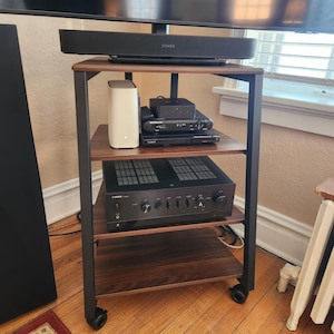 May include: A four-tier media stand with a dark wood grain finish and a black metal frame on wheels. The top shelf holds a black Sonos soundbar. The middle shelf contains a white router, a black streaming device, and a black Sony media player.