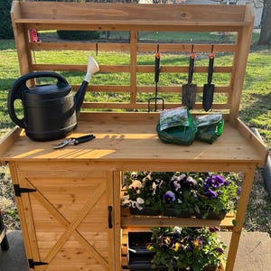 May include: A wooden potting bench with a slatted back panel, a single cabinet door with an X-brace design, and two open shelves. The bench holds a black plastic watering can, metal hand tools, two green plastic bags, and trays of colorful pansies.