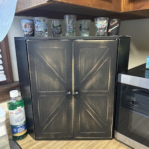 May include: A black wooden cabinet with two doors featuring a distressed finish and diagonal paneling, topped with seven souvenir shot glasses from various locations including Charleston, Colorado, and Nashville, arranged in a single row.